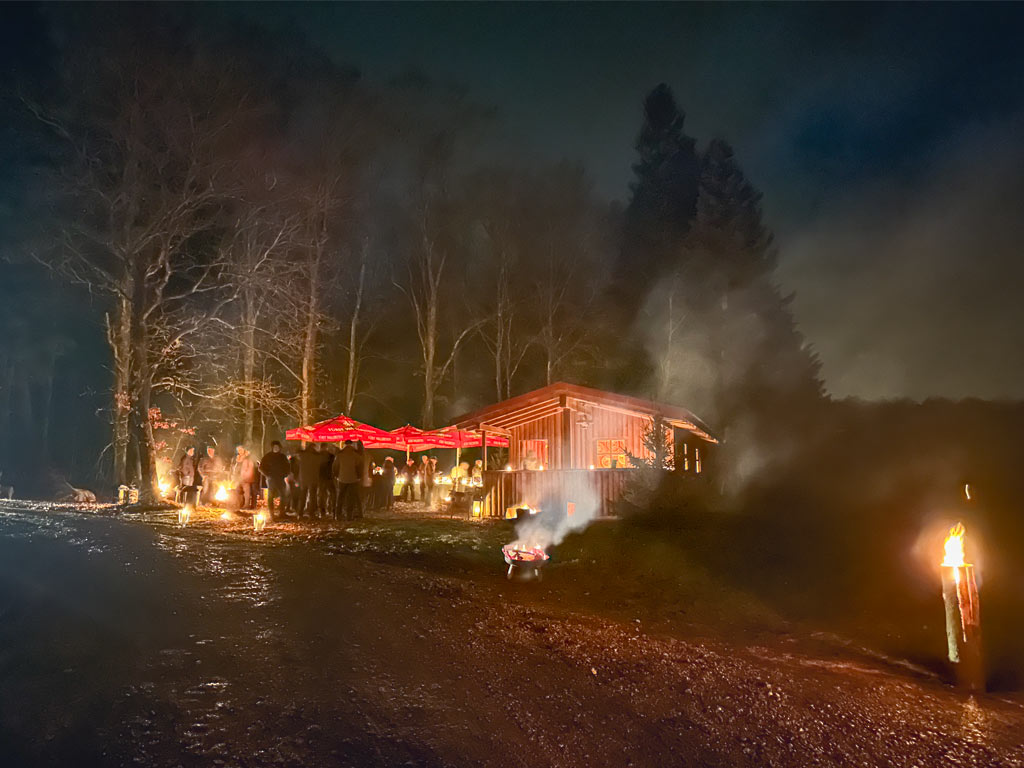 Menschen stehen unter roten Sonnenschirmen vor einer Jagdhütte im Wald bei Nacht